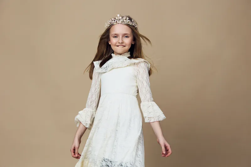 Joyful young girl in a white dress and crown, exuding confidence and playfulness against a beige background, perfect for concept of childhood royalty