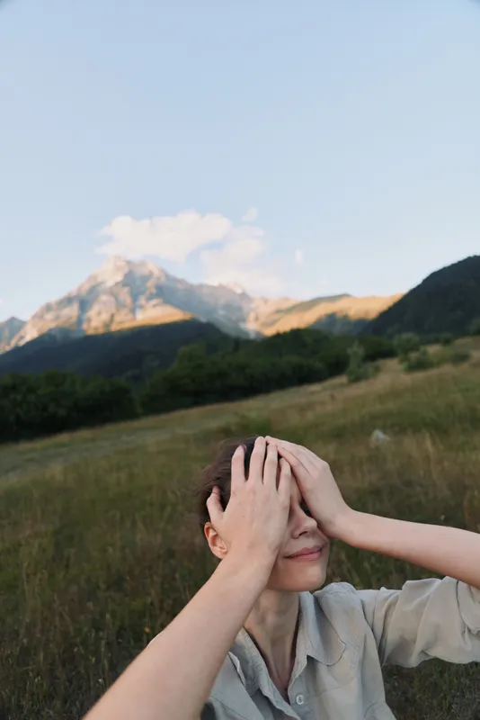 Woman in a light shirt stands in a grassy field with mountains in the background, hands covering her face, conveying stress and fatigue in an outdoor lifestyle moment