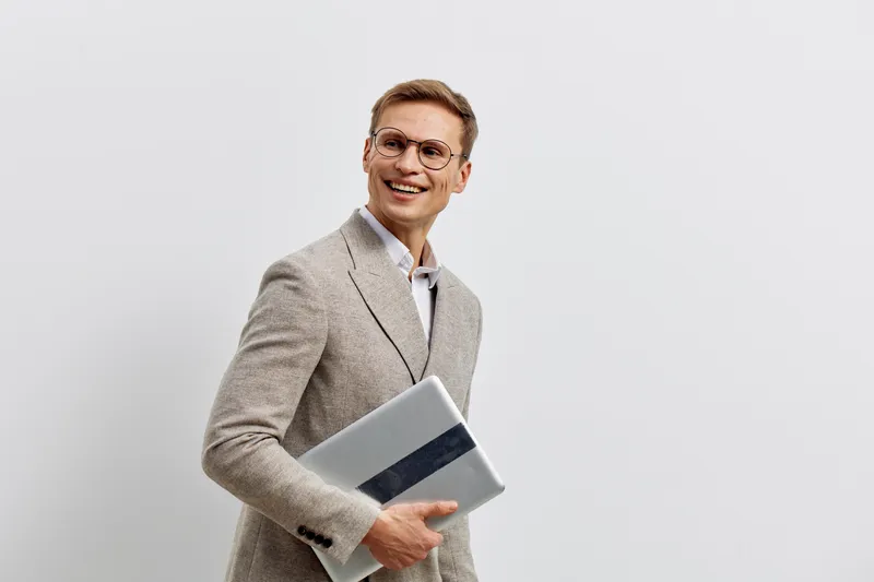 Confident smiling professional man in beige checked suit holding laptop and documents on plain white background. Business casual style, male, positive expression, office worker, corporate, success con