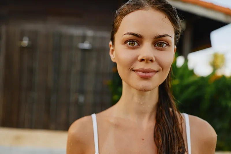 Young woman with wet hair smiles gently at camera outdoors near wooden building in natural light enjoying relaxed lifestyle and summer day with positive mood and casual appearance
