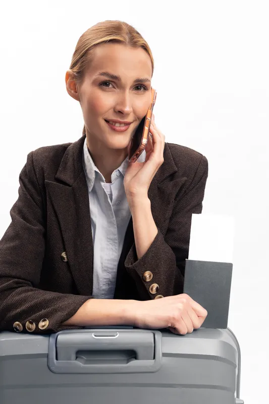 Smiling woman in business attire holds passport and smartphone while leaning on suitcase. White background studio shot. Concept of travel planning, business trip, and communication.