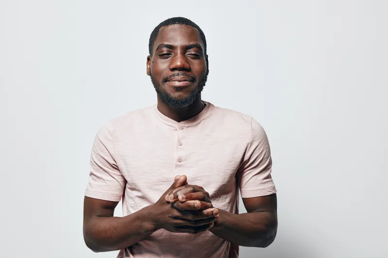 African American man with dark skin and beard wearing light pink shirt standing with hands clasped and eyes closed against white background in studio portrait.