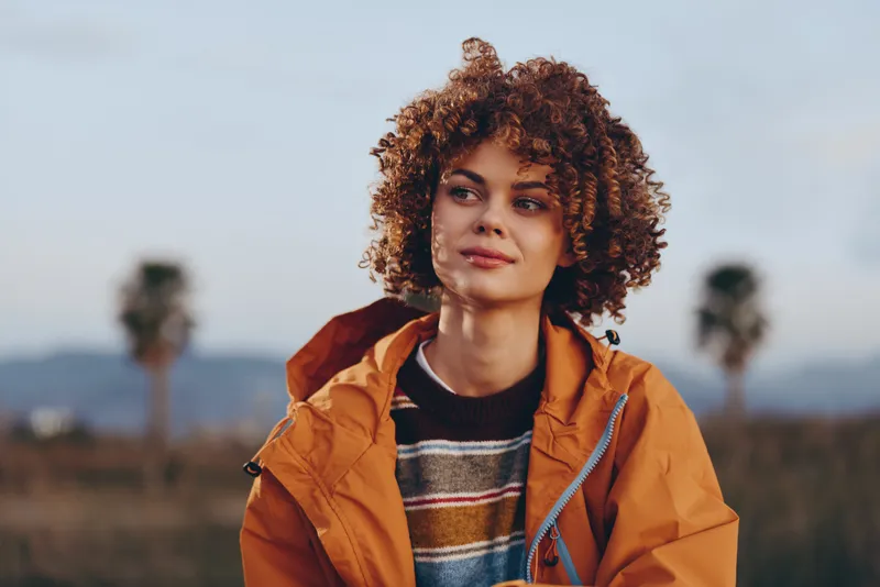 Woman curly hair portrait outdoors lifestyle jacket nature, sunset light, wearing striped sweater and orange jacket, looking at camera with calm expression, blurred mountains behind
