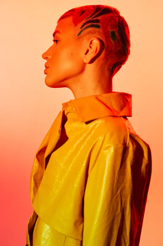 Woman with short bright red hair and shaved patterns wearing a yellow coat, standing in profile against an orange gradient background, showcasing bold fashion style.