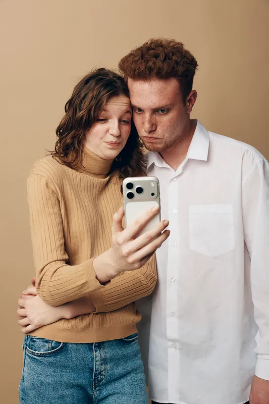 Young couple takes a selfie with a smartphone, showing playful and serious facial expressions. Casual clothing and neutral background create a relaxed, intimate atmosphere.