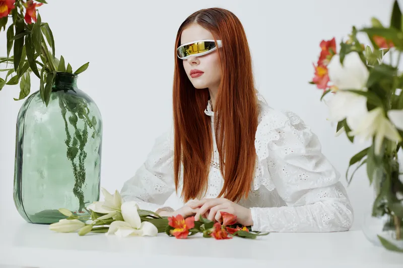 Stylish young woman with long red hair wearing futuristic sunglasses, surrounded by flowers and greenery in a bright, modern setting