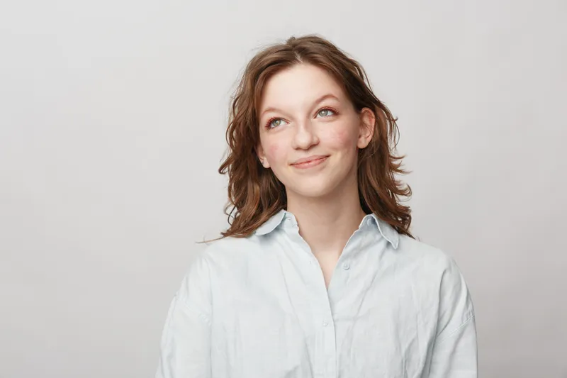 Young woman with wavy brown hair wearing white shirt looks up thoughtfully on light background. Concept of contemplation, hope, and casual lifestyle portrait with natural expression.