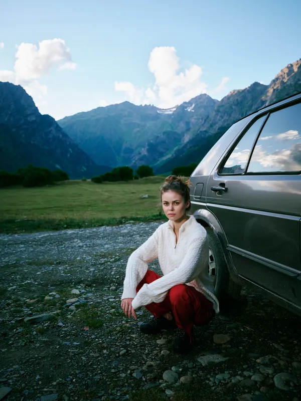 Woman beside a parked car in a rugged alpine landscape, mountains in the distance, casual sweater and jeans, capturing a calm outdoor moment of travel, nature, solitude scenery