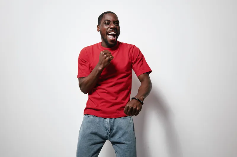 African American man with dark skin wearing red t shirt and jeans smiling and celebrating with fist pump against white background in studio.