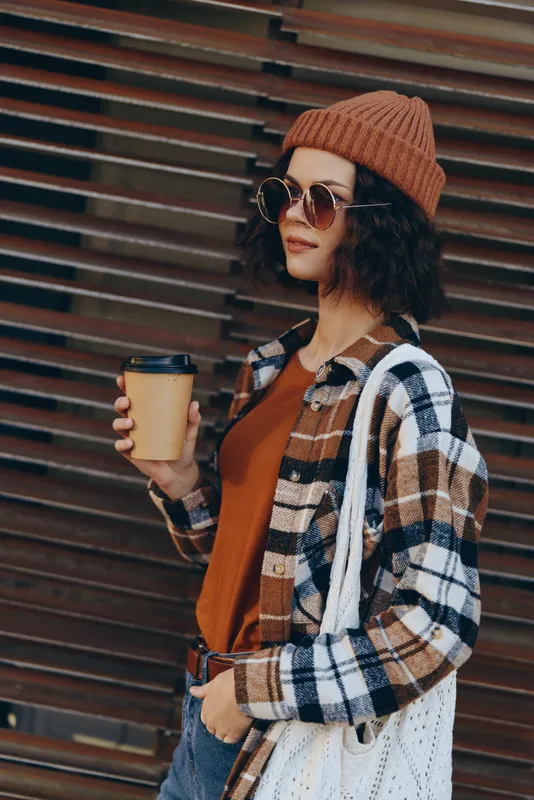 Woman coffee street casual beanie sunglasses plaid autumn tones, holding takeaway cup against urban wall. Authenticity, candid lifestyle, golden hour glow, mindful living and emotional storytelling