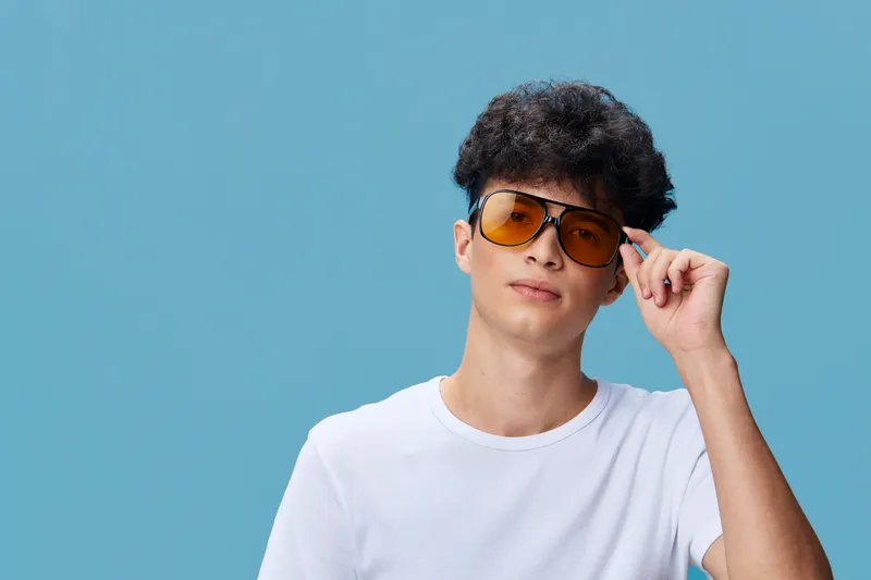 Young man wearing oversized yellow sunglasses touching frames against plain blue background, casual white t-shirt, stylish hairstyle, relaxed expression, modern look.