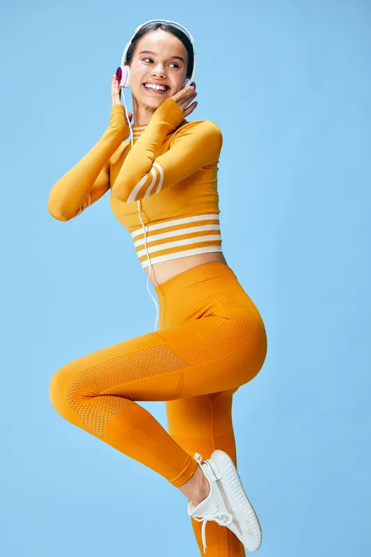 Woman fitness enthusiast wearing bright orange athleticwear with headphones, mid-air jump against a blue studio backdrop, exuding energy, movement, and athletic lifestyle.