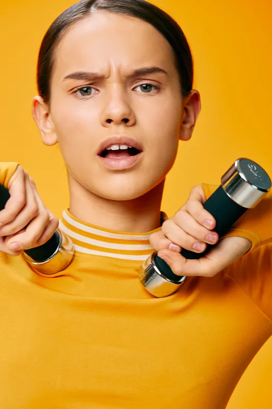 Fitness woman in an orange shirt lifts dumbbells with a focused expression, showcasing strength training, endurance and gym-style workout in a bold studio.