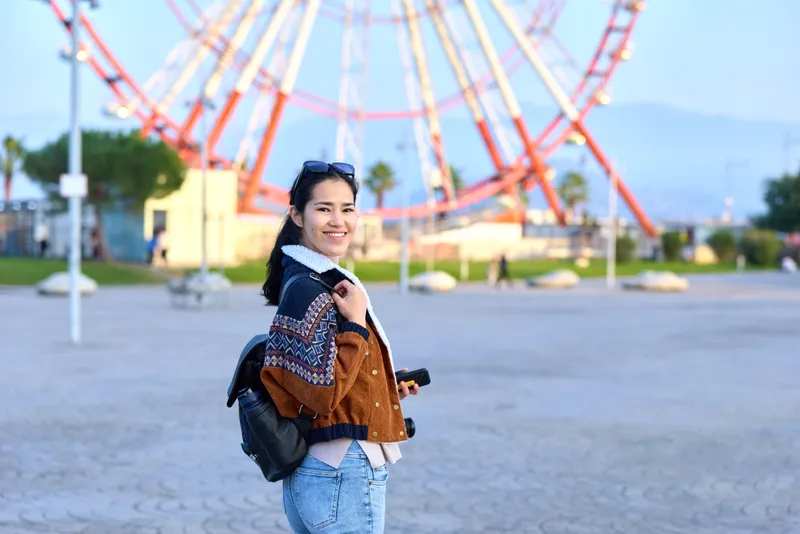 Woman traveler with backpack stands in an outdoor amusement park, smiling near a ferris wheel, casual fashion and sunny day mood, capturing a moment of travel and exploration