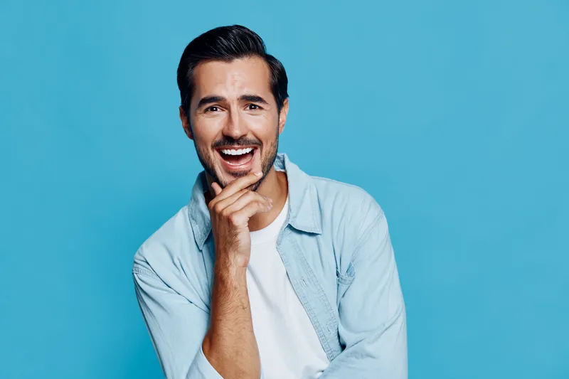 Smiling man with a charming expression, wearing a light blue shirt, looking confidently at the camera against a solid turquoise background. People lifestyle concept.