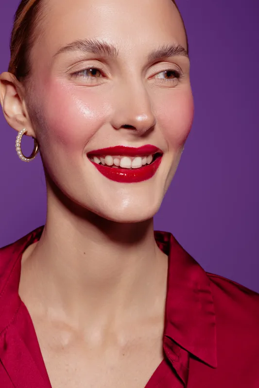 Close-up of smiling woman with red lipstick and pearl earring wearing red blouse on purple background. Concept of beauty, makeup, fashion, and confident female portrait.