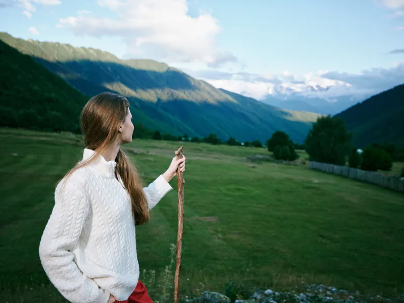 Landscape of rolling valley and distant mountains with a woman in a white sweater holding a walking stick, enjoying open countryside and fresh alpine air in nature