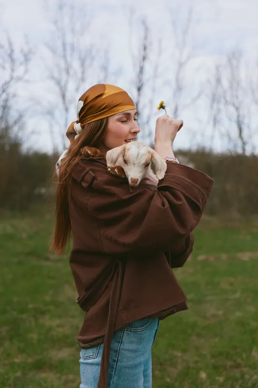 A lifestyle portrait of a woman outdoors embracing a dog in a grassy field, wearing a brown sweater and headscarf, capturing affection, nature, and companionship.