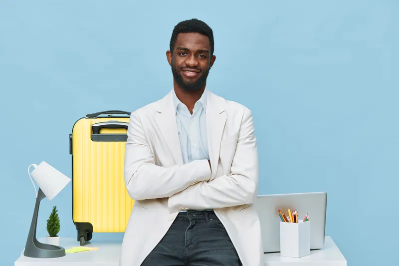 Confident smiling man with dark skin, curly hair, in light blazer and shirt, standing with arms crossed near desk with laptop, yellow suitcase, and office supplies against plain blue background, studi