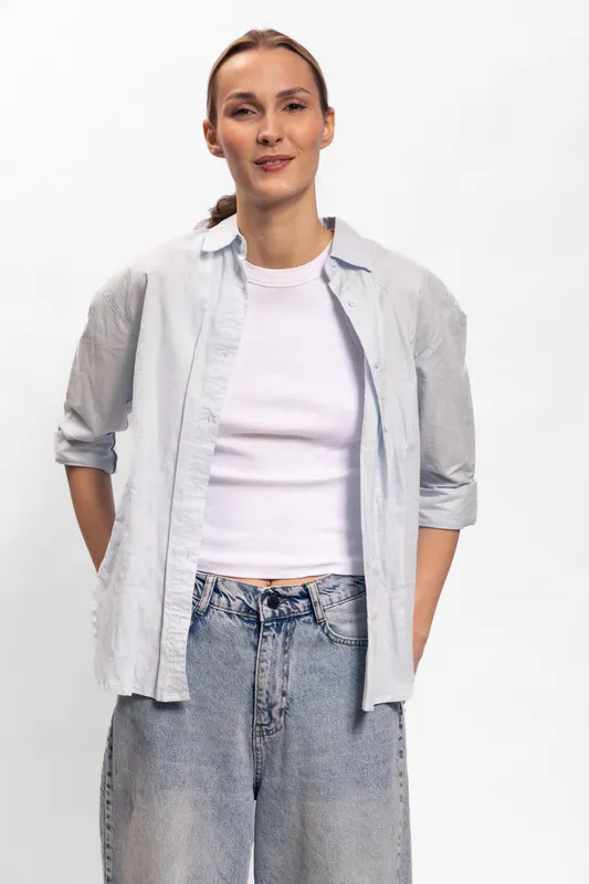 Confident woman in casual light blue shirt and white top stands with hands behind back on white background. Relaxed posture and neutral expression create calm, natural portrait concept.