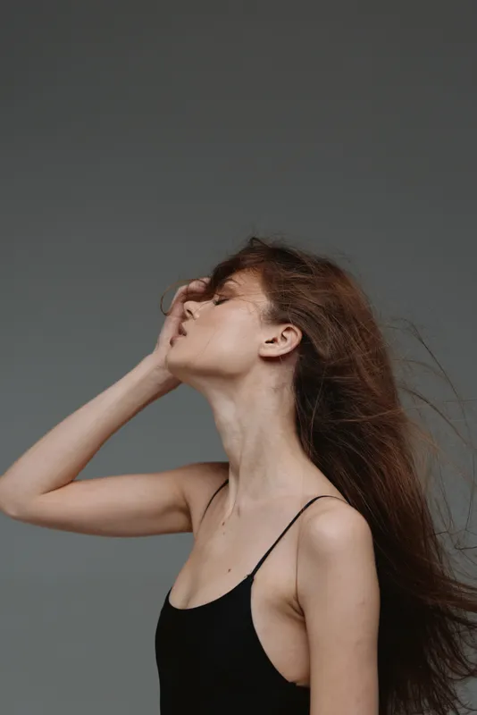 Profile of a young woman with long hair in black top posing gracefully against gray background.