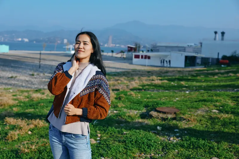 woman scarf outdoor field coast casual sweater shoreline landscape sunlight breeze, a calm image of a woman standing in a grassy area near the sea, open sky and distant hills add tranquil mood