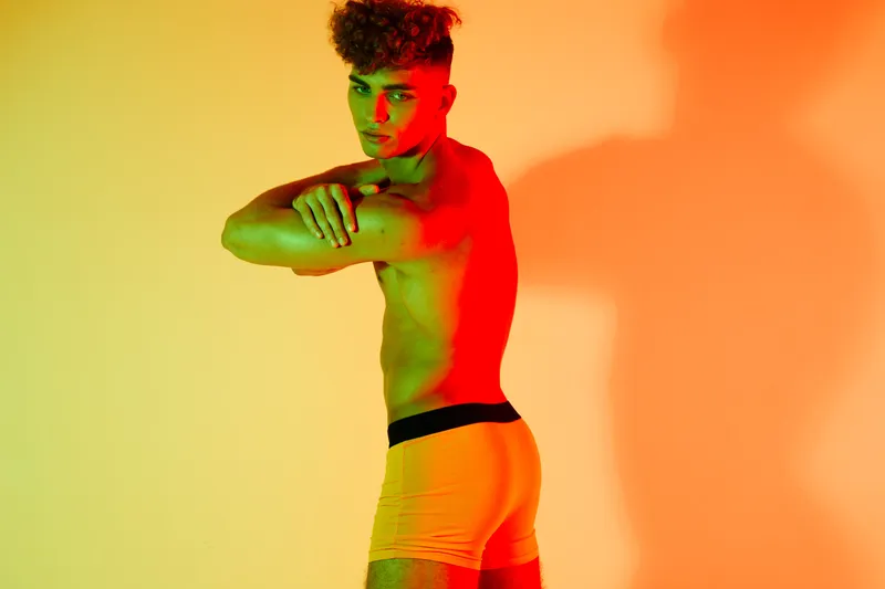 Man standing posing with crossed arms and bare torso wearing orange shorts in colorful lighting studio. Portrait of athletic young male fashion model showing muscle in vibrant indoor environment.
