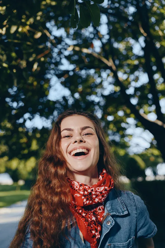 smiling woman with long hair outdoors wearing a red bandana and denim jacket under green trees on a sunny day
