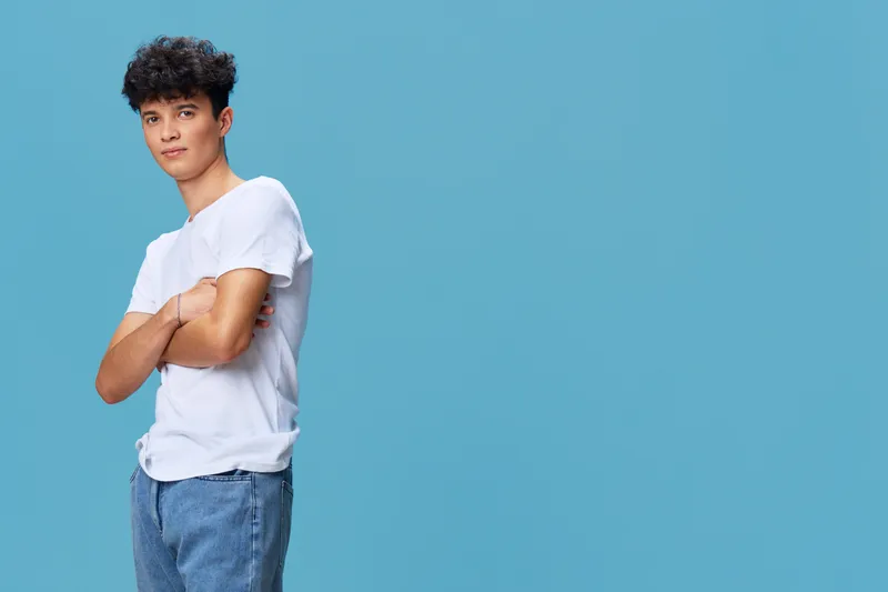 young man standing with crossed arms wearing casual white t-shirt and blue jeans against blue background showing relaxed confident attitude
