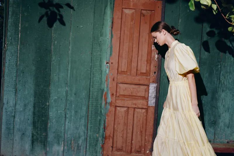 Woman in a pale yellow dress walks along a rustic doorway beside a green wall, creating a moody vintage street scene with soft light and quiet atmosphere.
