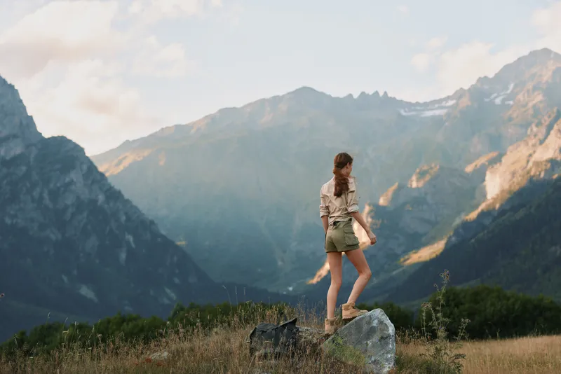 Young woman hiking in a rugged mountain landscape stands on a rock wearing a beige shirt and green shorts, feeling adventurous and focused