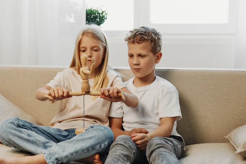 Two focused children play with a wooden toy on a beige couch in a bright room. The girl and boy concentrate on balancing the toy, showing curiosity and teamwork in a casual home setting.