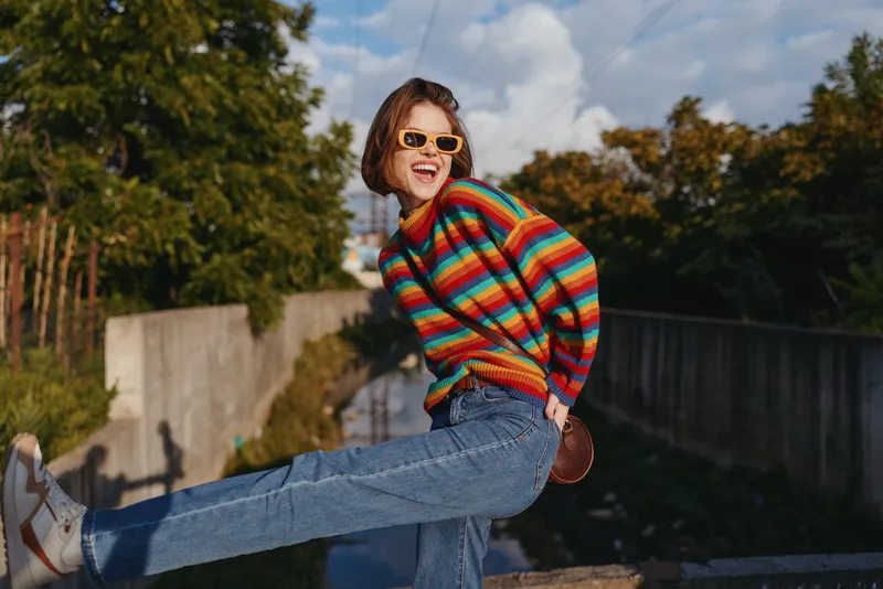 Woman in a colorful striped sweater and sunglasses, smiling and playful while kicking a leg on a sunny urban bridge. Casual outfit, jeans, short bob haircut, lifestyle travel.