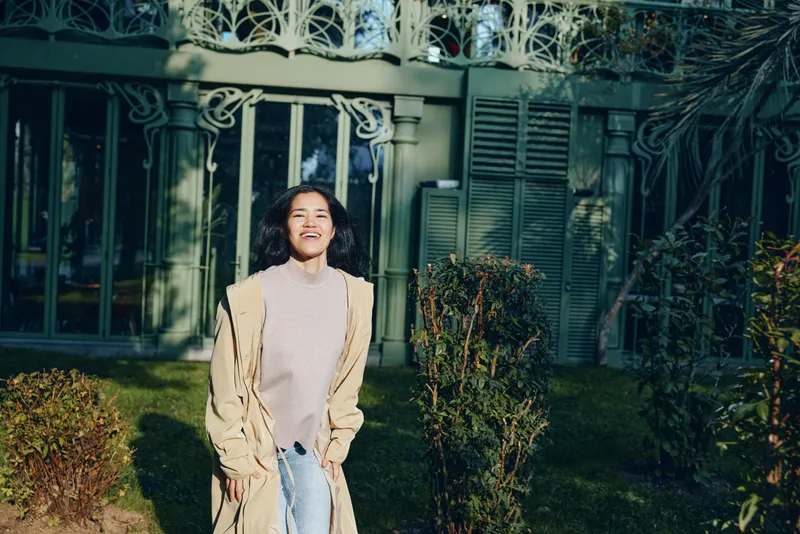 Woman in a beige coat stands outdoors in a garden beside a decorative gate, sunny day, casual fashion, candid smile, warm atmosphere, natural greenery and tranquil yard