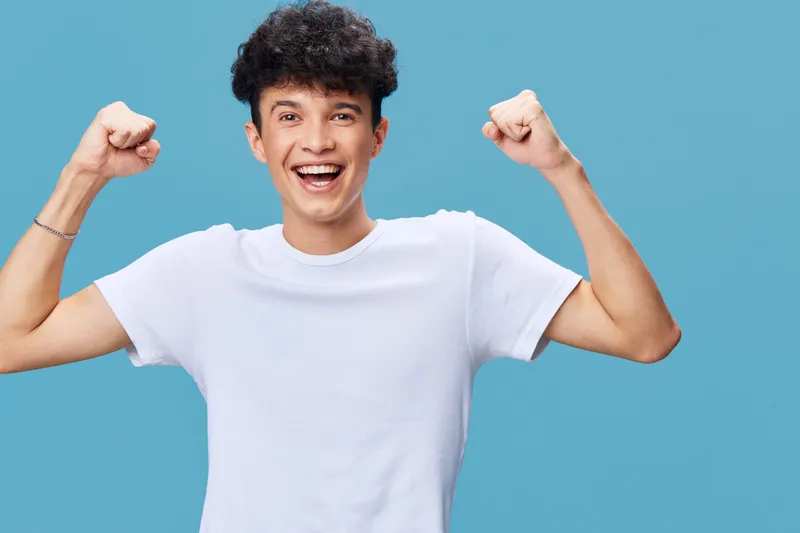 Smiling young man celebrating success with raised fists wearing a white t-shirt against a blue background, expressing happiness and energy.