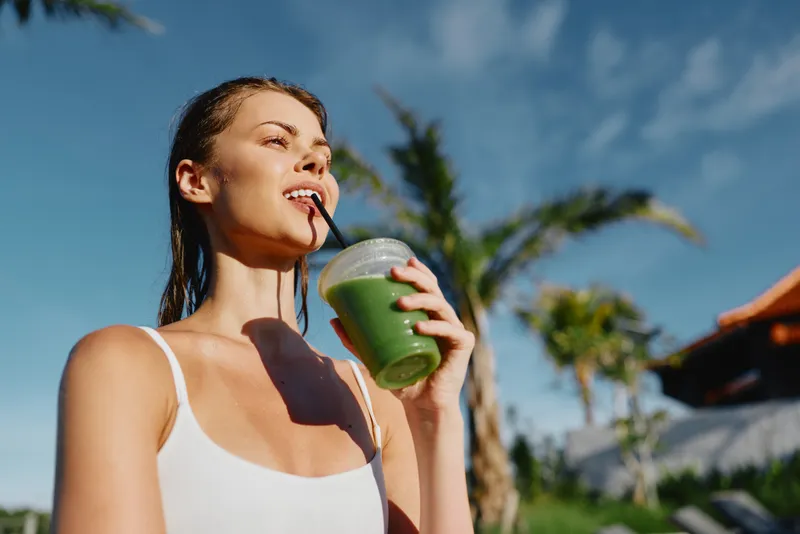 Cheerful woman outdoors, sipping a green smoothie under sun, relaxed posture, breeze in hair, tropical backdrop, vibrant colors, healthy lifestyle moment, carefree summer break