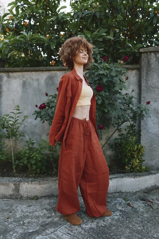 Young woman with curly hair wears a rust suit and beige top in a sunlit garden, standing relaxed beside a stone wall. Her pose conveys casual confidence and contemporary fashion