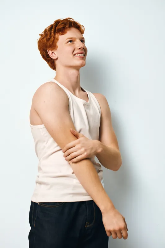 Young man with red hair in white tank top standing against white background looking to side with smile. Casual style, confident posture, and relaxed mood in studio lighting.