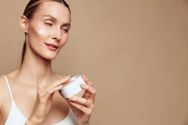 A young woman applies moisturizing cream on her cheek with a gentle smile, holding a white jar in a beige studio. Concept for skincare, beauty routine, and facial care.