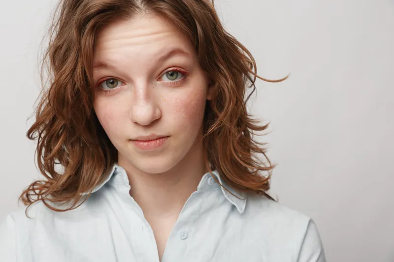 Young woman with wavy brown hair and green eyes looks directly at camera with neutral expression. She wears a light blue shirt against plain white background. Natural portrait concept.