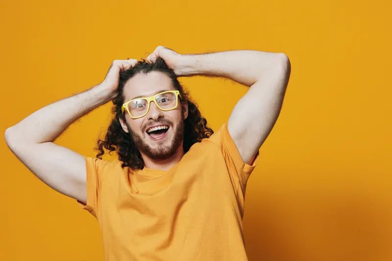 Happy young man with curly hair wearing yellow glasses and a bright orange shirt against a vibrant yellow background, expressing joy and excitement