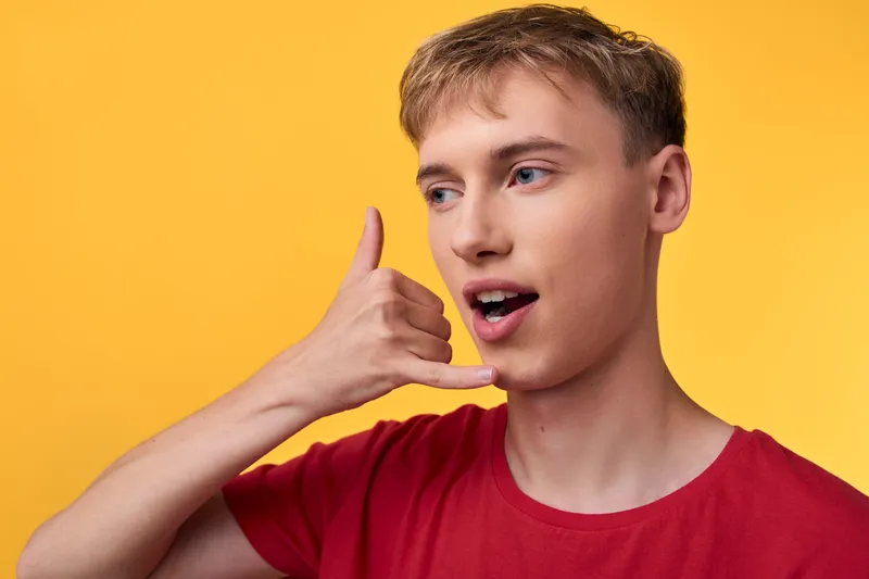 Young man in a red shirt makes a call gesture against a bright yellow background, creating a playful, casual vibe suitable for lifestyle, communication or social media visuals.