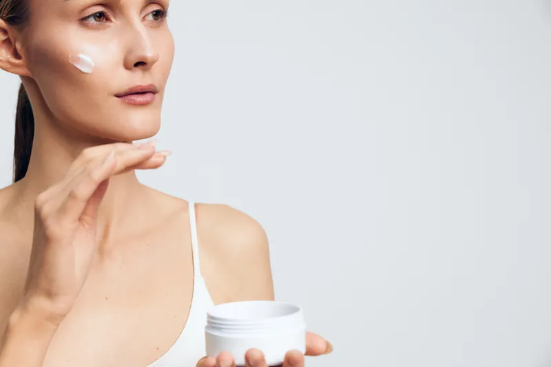 A woman applies moisturizer to her face in a bright, minimalist studio, capturing a focused skincare routine and a moment of self care and daily grooming.