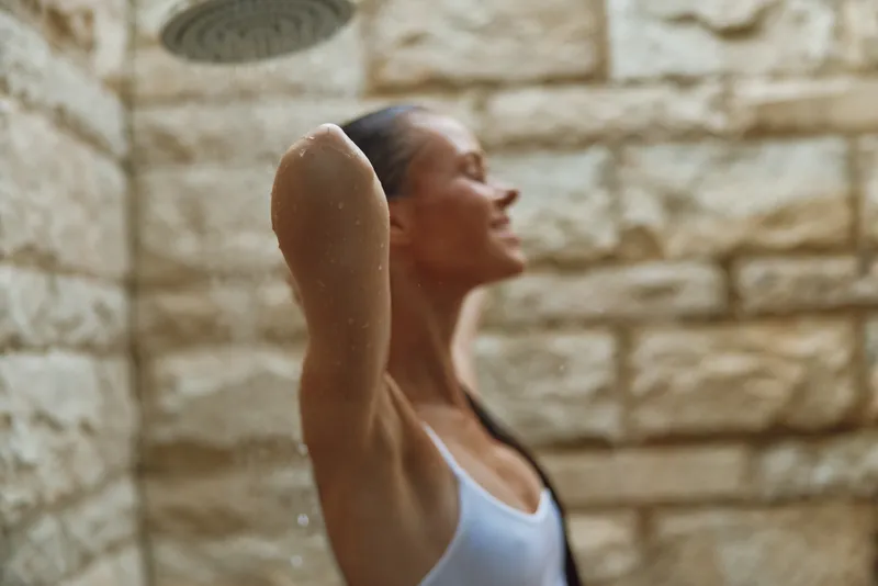 young woman enjoying outdoor shower with relaxed expression, wet hair, white swimsuit, stone wall background, natural light ambiance