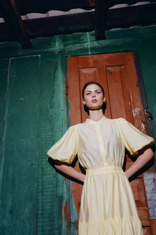 Fashion portrait of a woman in a pale yellow dress standing with hands on hips in front of a weathered green wall and wooden door, exuding bold vintage style.
