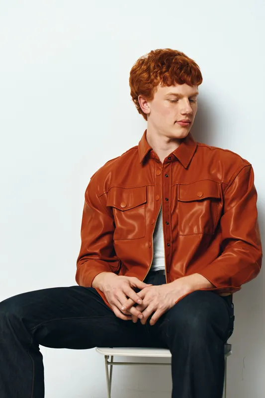 Young man with red hair sitting on white chair wearing brown leather jacket and black pants looking sideways against white background indoors casual fashion portrait.