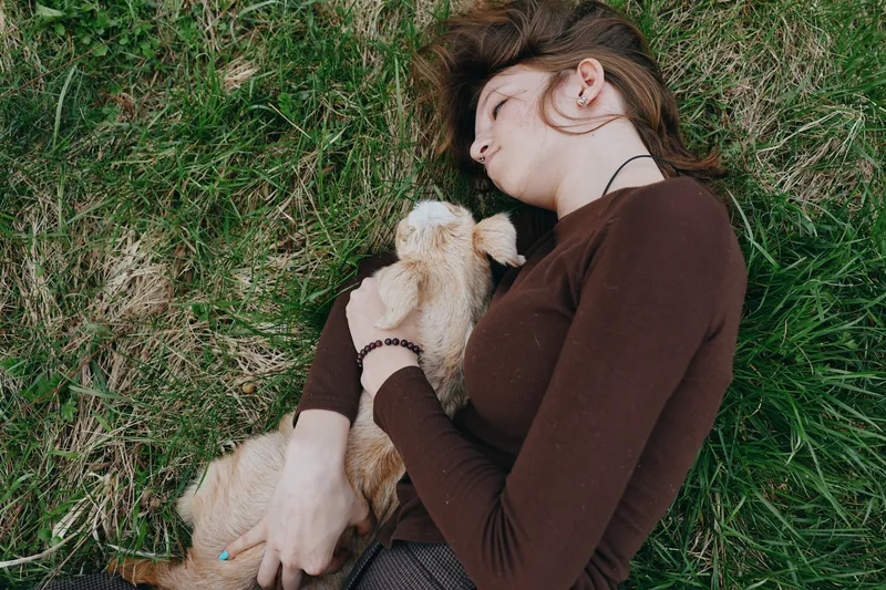 Relaxed woman lying on grass with a small dog, wearing a brown sweater, sharing a tender moment of companionship in nature. Soft daylight, calm mood, casual lifestyle, comfort, and affection outdoors.