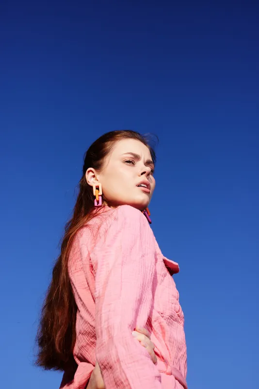 Young woman with long hair in a pink outfit against a clear blue sky, exuding confidence and style in a vibrant summer setting