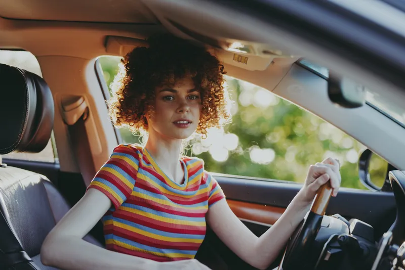 Woman driver with curly hair sits in colorful striped shirt inside a vintage car, capturing a casual lifestyle moment with retro film color and warm sunlight on the dashboard.