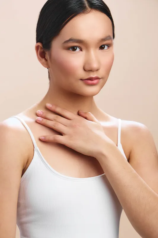 asian woman in a simple white tank top gazes softly at the camera, hand resting near her collarbone, creating an intimate studio portrait with warm lighting and natural calm.